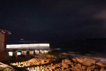 Sennen RNLI night This landscape photograph captures Sennen RNLI station on the coast of Sennen Cove in Cornwall at night during the early autumn season. The illuminated lifeboat slipway stands out against the dark sea, with coastal rocks visible in the foreground. The night sky is filled with faint stars, emphasizing the remote and tranquil atmosphere along this section of the Cornish coastline. The image highlights the coastal environment surrounding the RNLI station at Sennen Cove, providing a clear view of the structure’s proximity to the Atlantic and its vital position in this part of Cornwall.